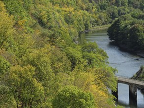 A quiet river flows under a small bridge, surrounded by dense forest with autumn leaves, Waldeck,