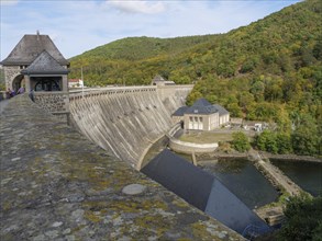 A large dam with a river and surrounding buildings, nestled in wooded hills under a blue sky,