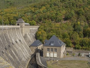 A massive dam with buildings and a dense, autumn-coloured forest landscape in the background,