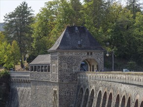 A dam with a historic stone building, surrounded by forest and trees, with a road and bridge lamps,