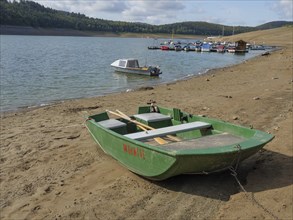 Small green boat on the sandy shore of a lake, other boats and hills in the background, Waldeck,