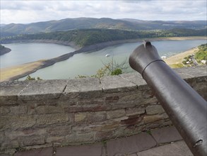 View from a stone wall with a cannon onto a lake surrounded by wooded hills under a cloudy sky,