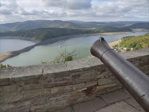 View from a stone wall with a cannon onto a lake surrounded by wooded hills under a cloudy sky,