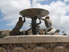 Bronze sculptures holding a large bowl of a fountain in a public square, valetta, mediterranean