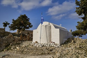 White chapel with Greek flag, surrounded by trees and sky with clouds, Kastro Monolithou, Chapel of