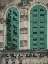 Two windows with green shutters on a stone building facade, slightly overgrown with plants,