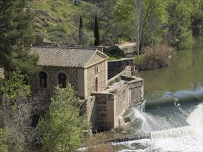 Small historic building on the riverbank with cascading waterfall, wrapped in a spring landscape,