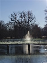Fountain in a wintry park, surrounded by bare trees and reflecting water under sunlight, ruurlo,