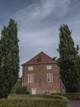 Historic brick house with gabled roof and windows, flanked by large green trees and hedges,