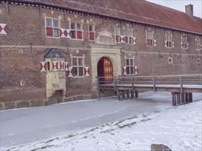 A historic brick building with colourful windows and a bridge over a frozen ditch on a winter's