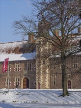 A historic castle with turrets, covered in snow and surrounded by trees with a waving flag,