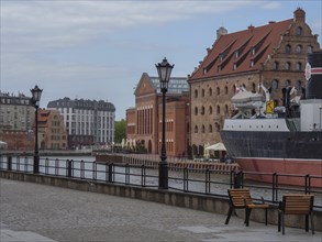 Riverbank with historic ship and buildings, street lamps and benches under a cloudy sky, Gdansk,