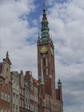 A large Gothic tower with a clock and historic house facades below, against a cloudy sky, Gdansk,
