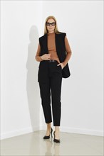 Serious woman in black vest, terracotta turtleneck, and breeches standing against a white office