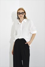 Indoor portrait of stylish blonde woman in white shirt with rolled-up sleeves and black trousers