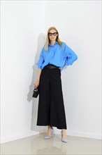 Stylish business woman in blue button down shirt and loose black skirt standing next to white wall