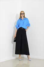 Confident woman in blue button down shirt and black maxi skirt posing over white wall