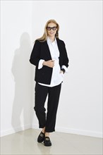 Trendy positive woman in cotton shirt, jacket and trousers walking in bright room with cheerful