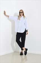 Woman in white cotton shirt and black trousers leans her hand against the wall while standing in a