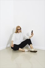 Cute woman wearing casual outfit read chat on smartphone sit on the floor against white wall