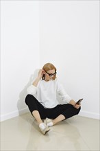 Cheerful woman in sweater and trousers sits on a floor with smartphone in her hand