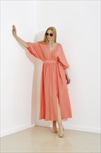 A stylish blonde woman in a coral maxi dress with a v-neckline leaning against white wall