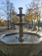 The water fountain, surrounded by autumn trees in the sunshine, creates a peaceful atmosphere,