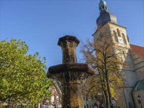 Water gushing through fountain and church tower with autumnal deciduous trees and blue sky,
