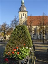 A historic church with a steeple in the background, surrounded by autumn trees and flowering plants