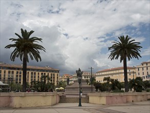 A large square with a statue in the centre, flanked by palm trees and surrounded by old buildings
