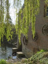 A brick building with large wooden wheels sits on the riverbank, surrounded by green trees and