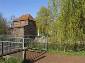 A narrow path with a bridge and metal railings leading to a brick building with a red roof,