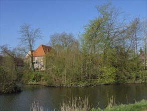 A quiet river flows beside a house with a red roof, surrounded by bare and partly green trees in a