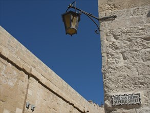 A decorative lamppost and street sign along an old stone wall under a blue sky, mdina,