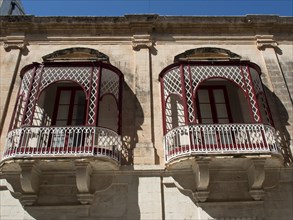 Two ornate and decorative iron balconies in front of a historic stone wall with windows, mdina,