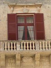 Close-up of a building with red shutters and a small balcony with opaque curtains, mdina,