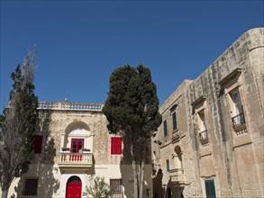 View of a square with historic buildings and red shutters, accompanied by trees under a cloudless