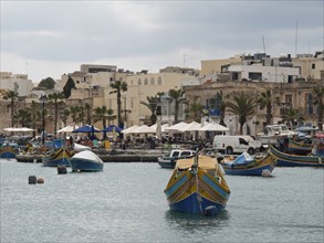 A row of fishing boats in front of a small town with palm trees, under a cloudy sky, marsaxlokk,