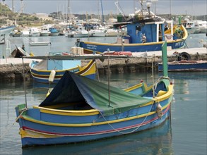 A blue and yellow fishing boat lies calmly in the water of a harbour, marsaxlokk, mediterranean