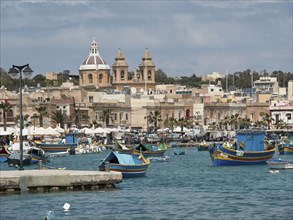 Boats in the harbour with an urban backdrop and a cathedral under a cloudy sky, marsaxlokk,