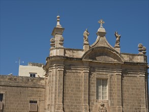 Stone church building with statues and cross on the roof, blue sky in the background, gozo,