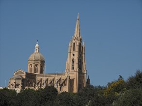 Gothic cathedral with high towers and a dome, surrounded by green vegetation under a clear sky,