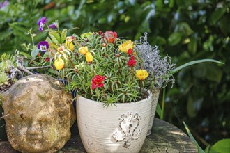 An antique-looking flower pot with colourful flowers next to a decorative bust in the garden,