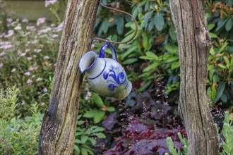 A decorative blue and white jug hangs from a rustic wooden frame in a flowery garden, Borken,