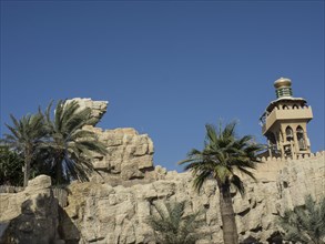 Traditionally designed buildings with palm trees and rock formations under a clear blue sky, Dubai,