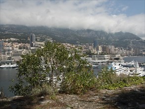 A view of a harbour with numerous yachts and boats, surrounded by mountains and a city under a