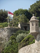 A historic watchtower with a flag in the background, surrounded by green vegetation and trees,