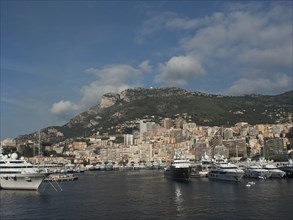 A harbour with several yachts and ships, flanked by mountains and a dense arrangement of buildings,