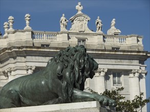 Bronze lion statue in front of a magnificent building with several stone figures under a clear sky,
