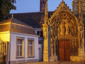 An illuminated gothic façade with detailed sculptures next to a historic building at dusk,